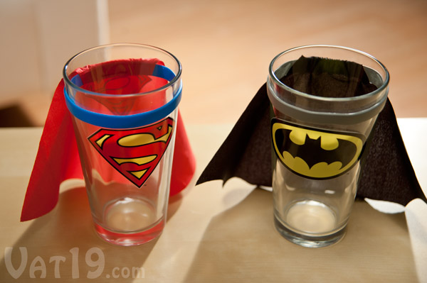 Superman and Batman Pint Glasses with capes sitting on a table.